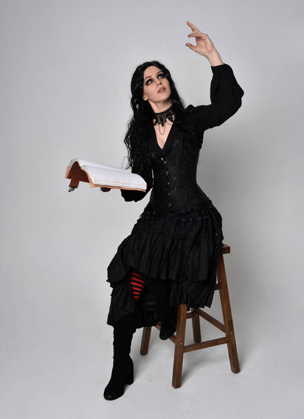 Full length portrait of dark haired woman wearing  black victorian witch costume.  sitting pose on a chair, with gestural hand movements,  against studio background. - Photo, Image