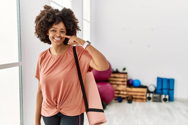 African american woman with afro hair holding yoga mat at pilates room pointing with hand finger to face and nose, smiling cheerful. beauty concept  - Photo, Image