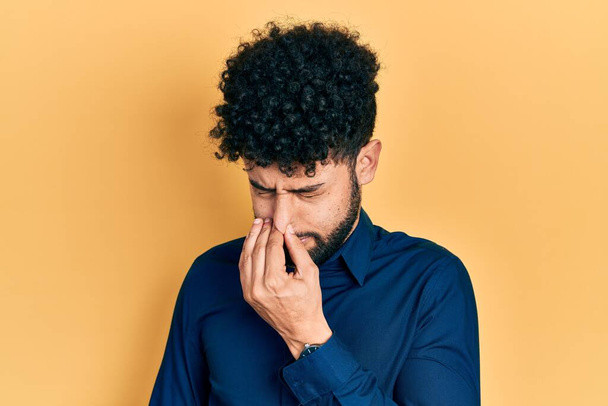 Young arab man with beard wearing casual shirt smelling something stinky and disgusting, intolerable smell, holding breath with fingers on nose. bad smell  - Фото, изображение