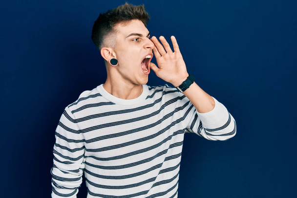 Young caucasian boy with ears dilation wearing casual striped shirt shouting and screaming loud to side with hand on mouth. communication concept.  - Photo, Image