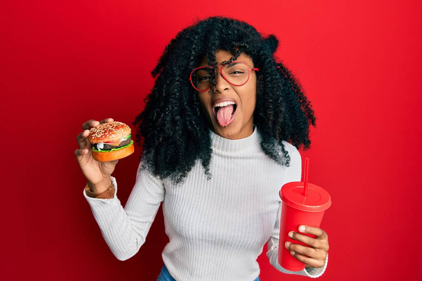 Afroamerikanerin mit Afro-Haaren isst einen leckeren klassischen Burger und Soda streckt die Zunge glücklich mit witzigem Ausdruck heraus.  - Foto, Bild
