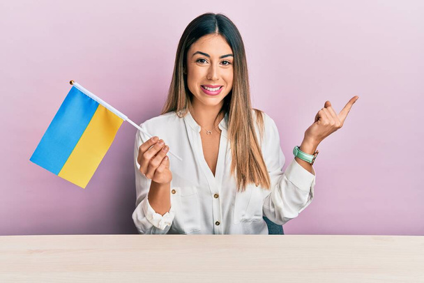 Jeune femme hispanique tenant drapeau ukraine assis sur la table souriant heureux pointant avec la main et le doigt sur le côté  - Photo, image