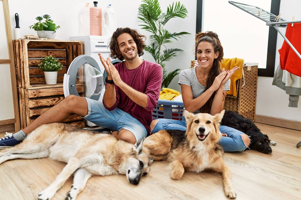 Молодая латиноамериканская пара стирает белье с собаками, хлопающими в ладоши и аплодирующими счастливыми и радостными, улыбающимися гордыми руками вместе  - Фото, изображение