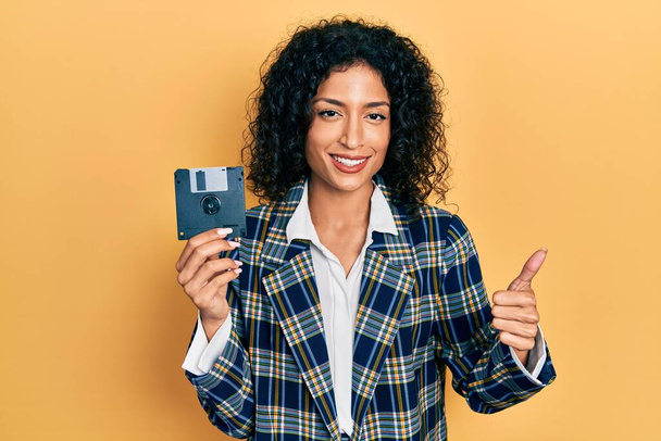 Young latin girl holding floppy disk smiling happy and positive, thumb up doing excellent and approval sign  - Foto, imagen
