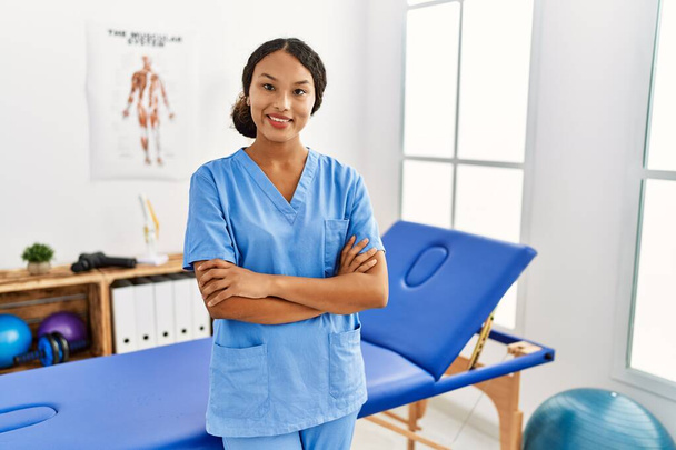Güzel İspanyol fizyoterapist kadın ağrıyı dindirme kliniğinde kollarını kavuşturup kameraya bakarken gülümsüyor. pozitif kişi.  - Fotoğraf, Görsel
