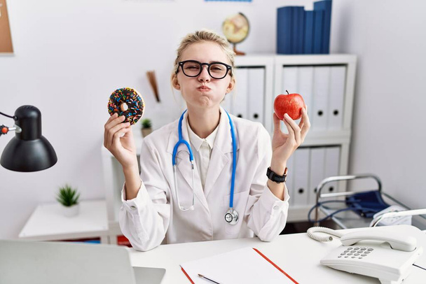 Junge Ärztin mit rotem Apfel und Donut in der Klinik, die mit lustigem Gesicht Wangen pustet. Mund mit Luft aufgeblasen, Luft einfangen.  - Foto, Bild