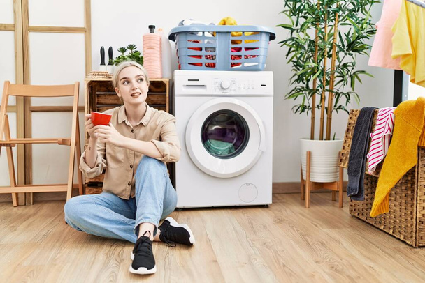 Joven mujer caucásica bebiendo café esperando la lavadora en la sala de lavandería - Foto, Imagen