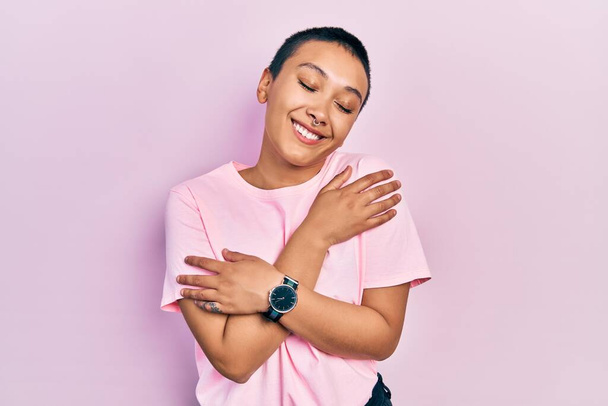 Beautiful hispanic woman with short hair wearing casual pink t shirt hugging oneself happy and positive, smiling confident. self love and self care  - Fotografie, Obrázek
