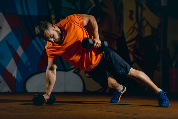 Full-length portrait of sportive man doing push-ups with dumbbells, training in sport gym - Фото, зображення