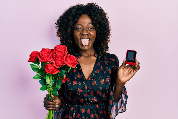 Young african american woman holding bouquet of flowers and engagement ring sticking tongue out happy with funny expression.  - Фото, зображення