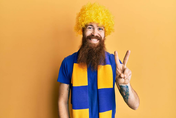 Kızıl saçlı, uzun sakallı futbol holigan amigoluk oyunu komik peruk takıyor ve parmaklarıyla iki numarayı gösterirken kendinden emin ve mutlu gülümsüyor..  - Fotoğraf, Görsel