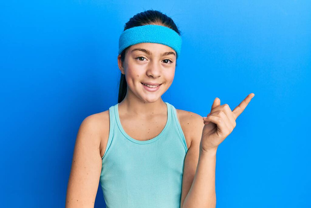 Όμορφη μελαχρινή κοριτσάκι φορώντας αθλητικά ρούχα και diadem χαμογελώντας χαρούμενος δείχνοντας με το χέρι και το δάχτυλο στο πλάι  - Φωτογραφία, εικόνα