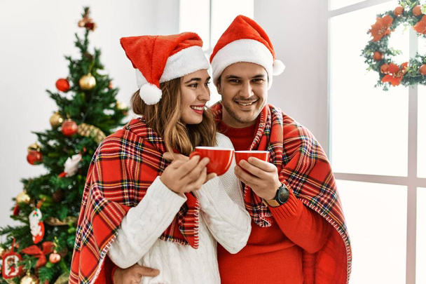 Jeune couple hispanique portant un chapeau de Noël et une couverture. Debout avec le sourire sur le visage boire du café à la maison. - Photo, image