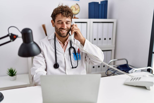 Молодой латиноамериканец в форме доктора разговаривает по телефону в клинике - Фото, изображение