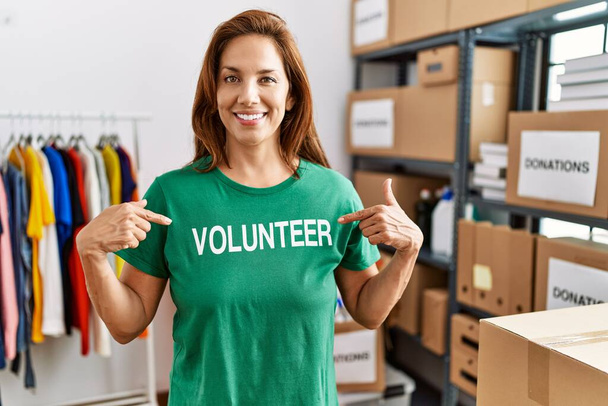 La donna ispanica di mezza età che indossa una t-shirt volontaria alle donazioni sta sorridendo felice indicando con mano e dito  - Foto, immagini