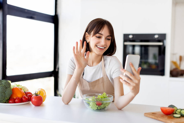 Video blog about healthy food. Happy young woman with bowl of fresh salad on table recording video on smartphone - Photo, Image