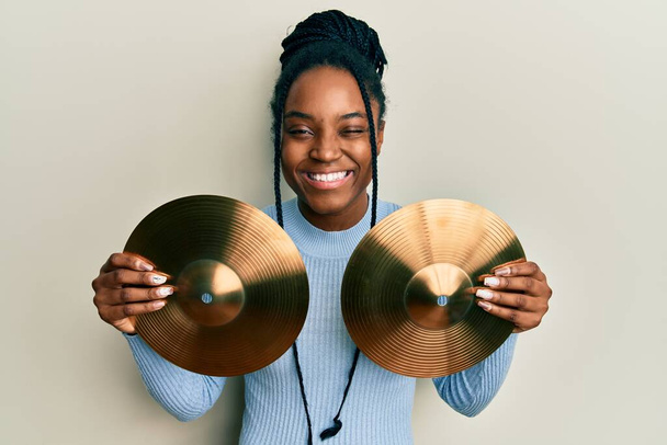 Femme afro-américaine aux cheveux tressés tenant des plaques de cymbales dorées clignant des yeux en regardant la caméra avec une expression sexy, gaie et heureuse visage.  - Photo, image