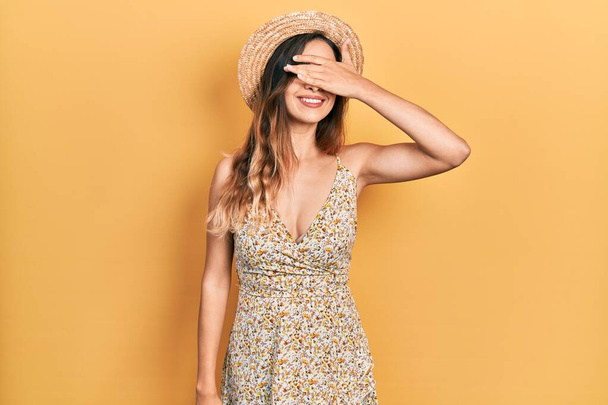 Young hispanic girl wearing summer hat smiling and laughing with hand on face covering eyes for surprise. blind concept.  - Fotoğraf, Görsel