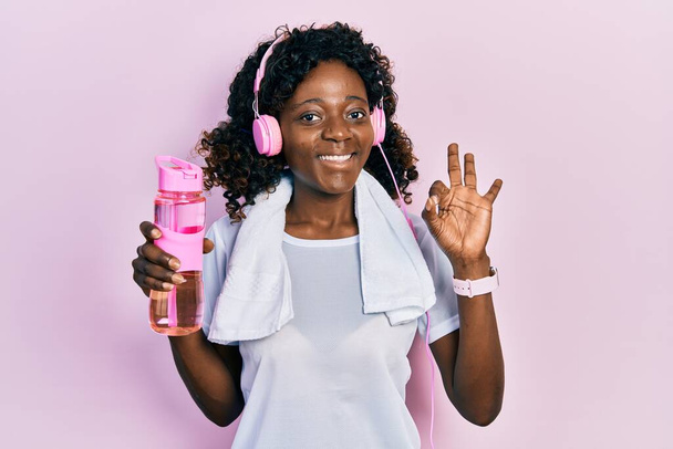 Young african american woman wearing sportswear holding water bottle using headphones doing ok sign with fingers, smiling friendly gesturing excellent symbol  - Foto, afbeelding