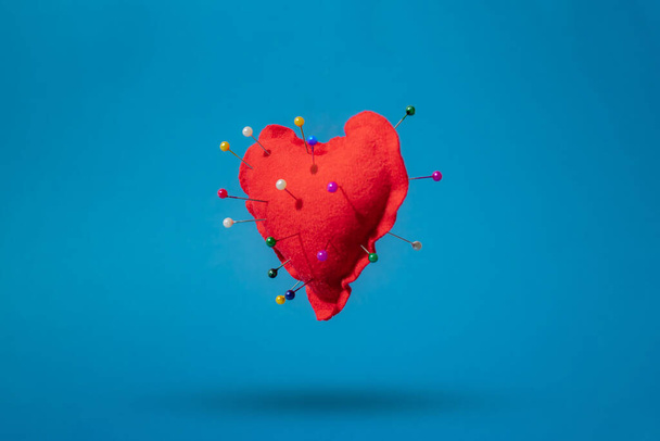 Een rood hart dat in de lucht zweeft. Gestoken met pinnen van verschillende kleuren. Blauwe achtergrond. - Foto, afbeelding