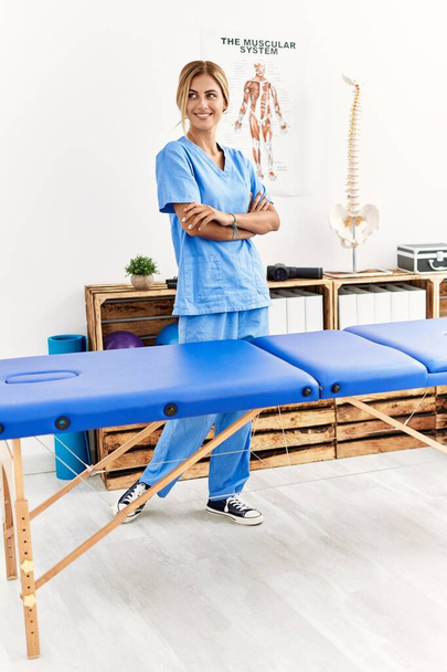 Nuori valkoihoinen fysio terapeutti hymyilee onnellinen seisoo kädet ristissä ele klinikalla - Valokuva, kuva