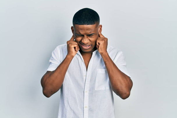 Jeune homme noir portant une chemise blanche décontractée couvrant les oreilles avec des doigts avec une expression agacée pour le bruit de la musique forte. concept sourd.  - Photo, image