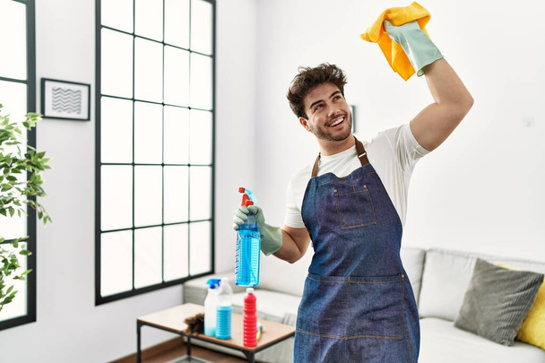 Genç İspanyol adam evde dans ediyor, temizlik spreyi ve bez tutuyor. - Fotoğraf, Görsel