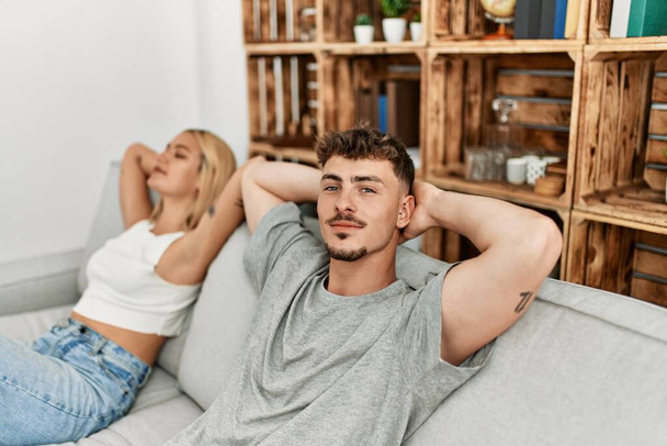 Mladý kavkazský pár relaxovat s rukama na hlavě sedí na pohovce doma. - Fotografie, Obrázek
