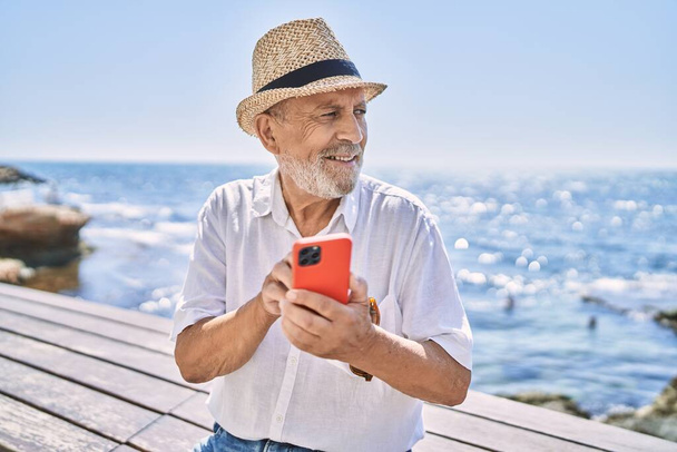 Hombre mayor sonriendo confiado usando sombrero de verano usando teléfono inteligente en la playa - Foto, Imagen