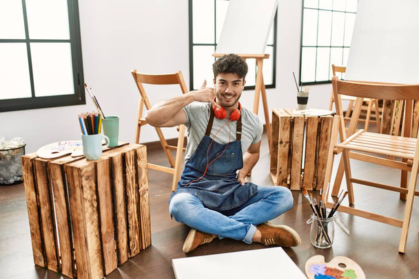 Young hispanic man sitting at art studio smiling doing phone gesture with hand and fingers like talking on the telephone. communicating concepts.  - 写真・画像