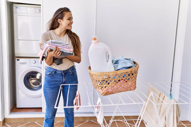 Young hispanic girl doing laundry holding folded clothes at home. - Foto, Imagem