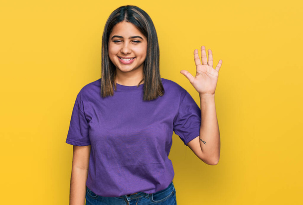 Joven chica hispana vistiendo casual camiseta púrpura mostrando y señalando hacia arriba con los dedos número cinco mientras sonríe confiado y feliz.  - Foto, imagen