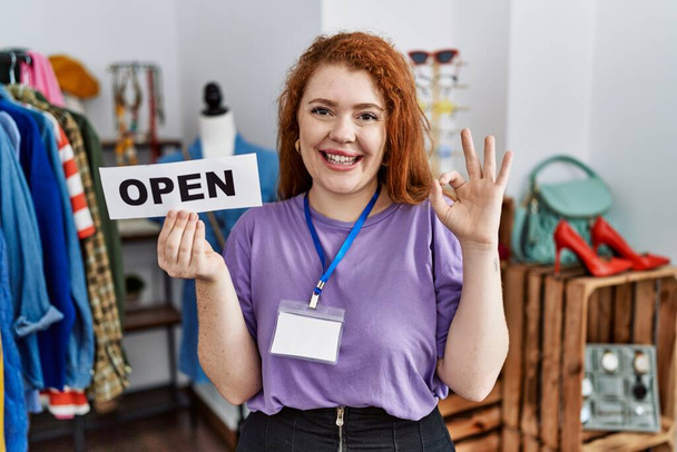 Młoda ruda kobieta trzyma baner z otwartym tekstem w sklepie robi ok znak palcami, uśmiechnięty przyjazny gest doskonały symbol  - Zdjęcie, obraz