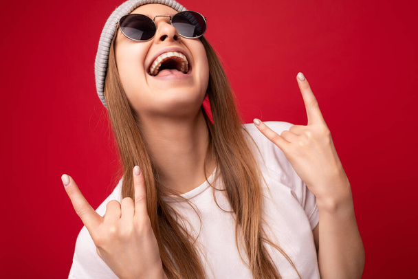 Primer plano de la joven emocional positiva feliz atractiva mujer rubia oscura con emociones sinceras con camiseta blanca casual, sombrero gris y gafas de sol aisladas en el fondo rojo con espacio de copia y - Foto, imagen