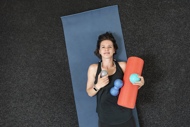 Das junge athletische Mädchen liegt auf einer Yogamatte mit verschiedenen myofaszialen Rollen. Ausrüstung für MFR. Myofasziale Freisetzung. Ansicht von oben. - Foto, Bild