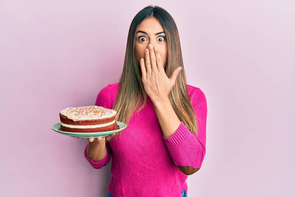Giovane donna ispanica che tiene la torta di carote che copre la bocca con la mano, scioccata e spaventata per errore. espressione sorpresa  - Foto, immagini