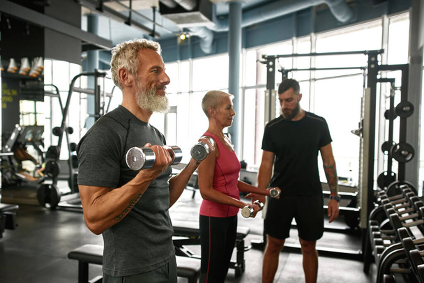 Approachable trainer supporting elderly couple while gym workout - Foto, Imagen