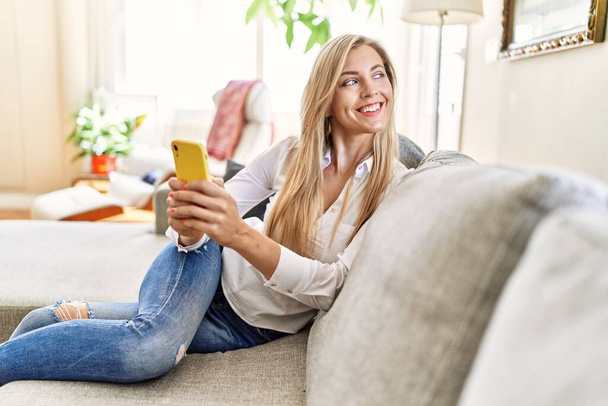 Jovem loira usando smartphone sentado no sofá em casa. - Foto, Imagem