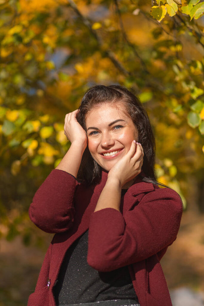 Portrait of a joyful young woman enjoying in the autumn park. Beautiful brunette girl in autumn red coat and black sweater. Relax in nature. High quality photo - Photo, image