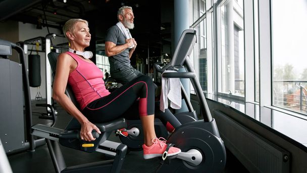 Couple of cheerful aged woman and man on gym cycles - Photo, image