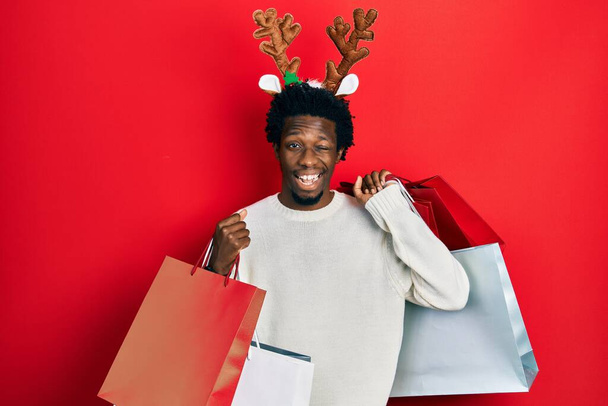 Young african american man wearing deer christmas hat holding shopping bags winking looking at the camera with sexy expression, cheerful and happy face.  - Photo, Image