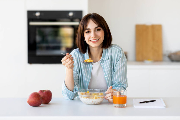 Terve aamiaiskonsepti. Onnellinen nainen syö mysli kulhoon omenat ja mehu, istuu moderni keittiö sisustus - Valokuva, kuva