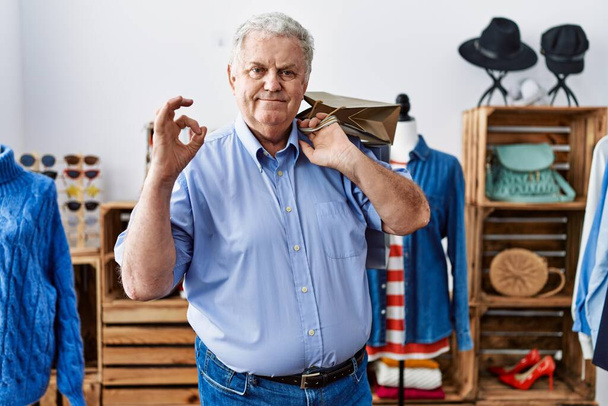 Starszy mężczyzna z siwymi włosami trzymający torby na zakupy w sklepie robi ok znak palcami, uśmiechając przyjazny gest doskonały symbol  - Zdjęcie, obraz
