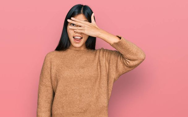 Gyönyörű fiatal ázsiai nő visel alkalmi téli pulóver kukucskál sokk borító arc és a szemek kézzel, nézi az ujjakon keresztül kínos kifejezés.  - Fotó, kép