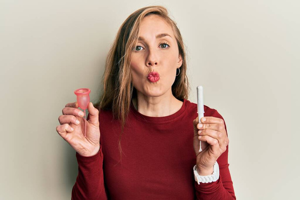 Young blonde woman holding menstrual cup and tampon looking at the camera blowing a kiss being lovely and sexy. love expression.  - Foto, Imagem