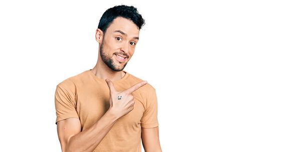 Hombre hispano con barba vistiendo camiseta casual alegre con una sonrisa en la cara apuntando con la mano y el dedo hacia un lado con expresión feliz y natural  - Foto, Imagen