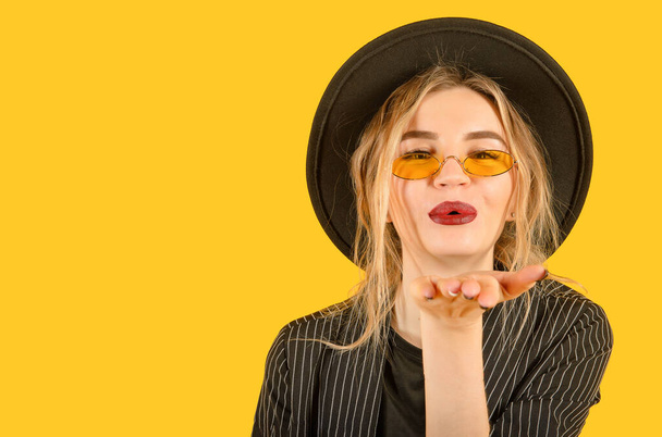 Young girl posing on yellow background direct look air kiss close up brown clothes yellow glasses copy space minimalistic - 写真・画像