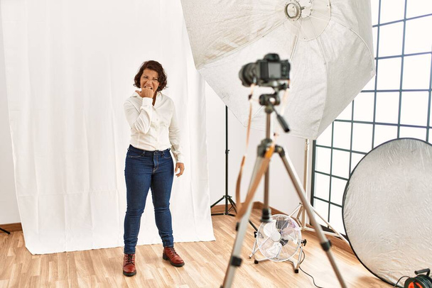 Middle age hispanic woman posing as model at photography studio looking stressed and nervous with hands on mouth biting nails. anxiety problem.  - Foto, immagini