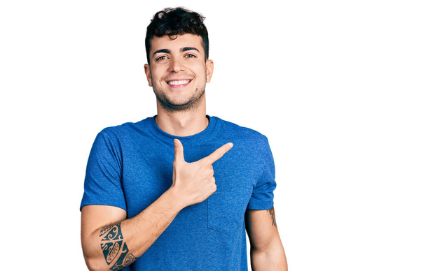 Joven hombre hispano vistiendo camiseta casual alegre con una sonrisa en la cara apuntando con la mano y el dedo hacia un lado con expresión feliz y natural  - Foto, imagen