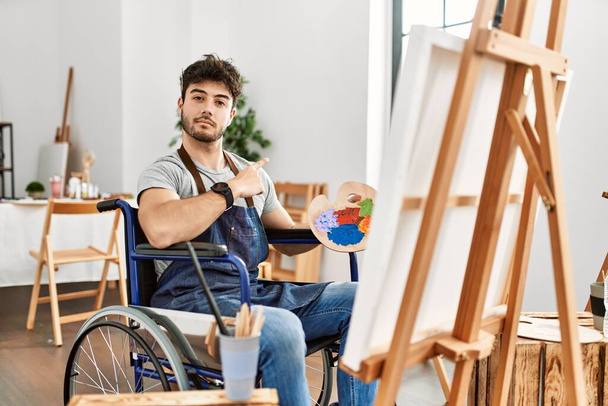 Junger hispanischer Mann sitzt im Rollstuhl im Kunstatelier und zeigt mit dem Finger zur Seite, zeigt Werbung, ernstes und ruhiges Gesicht  - Foto, Bild